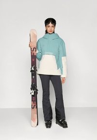 Lyžařský outfit zahrnující dvoubarevnou bundu modré a bílé barvy, černé kalhoty, černé boty a růžové lyže s vázáním; obsahuje lyžařské brýle.