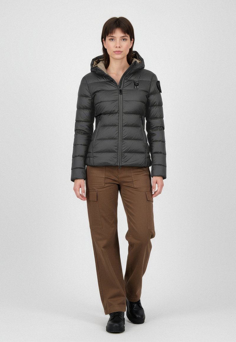 Dunkelgraue Steppjacke mit taillierter Silhouette und Kapuze, ausgestattet mit einem Reißverschluss und dezentem Branding. Kombiniert mit braunen Cargo-Hosen und schwarzen Stiefeln.