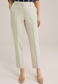 Pantalons beiges ajustés avec une coupe droite, des plis à l'avant et des poches latérales, associés à des chaussures à talons hauts nude. Texture de tissu lisse.