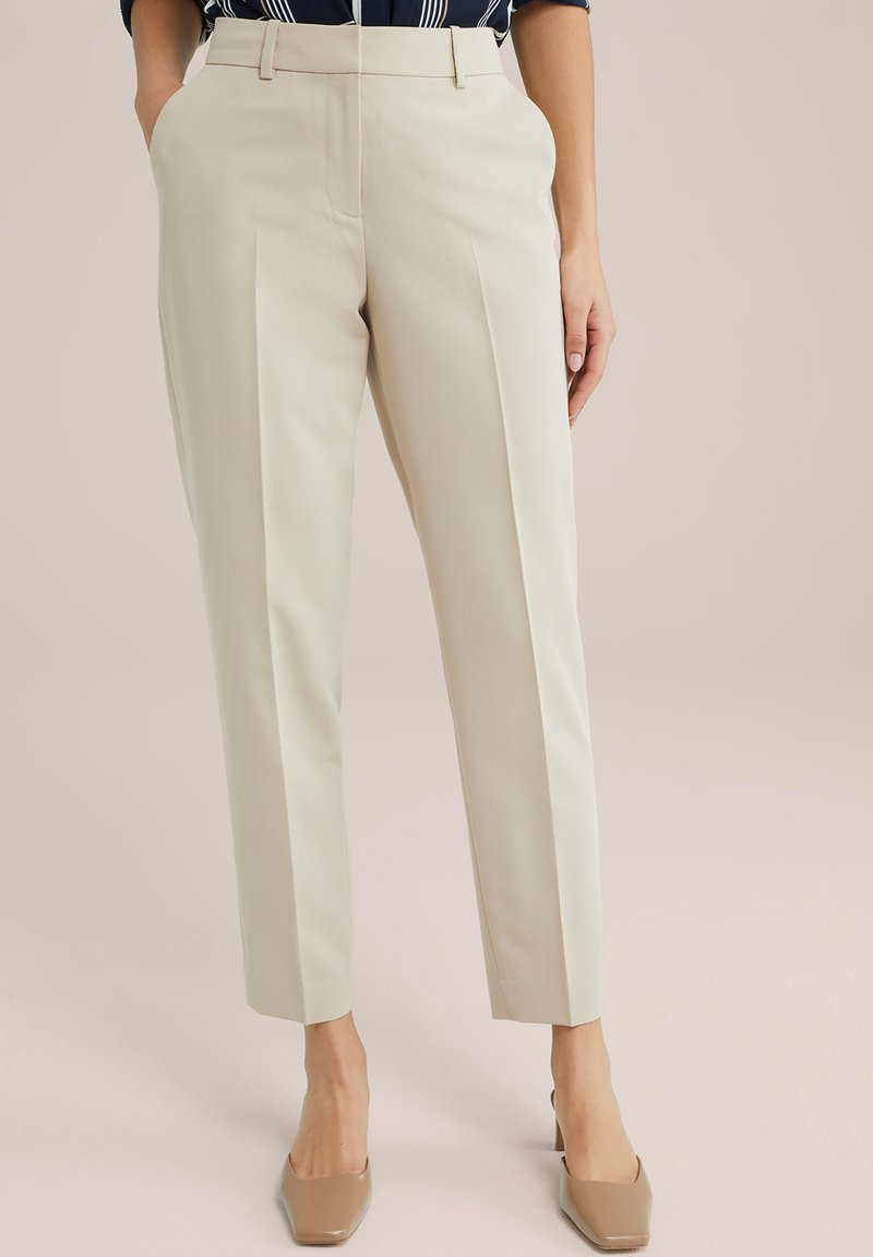 Pantalons beiges ajustés avec une coupe droite, des plis à l'avant et des poches latérales, associés à des chaussures à talons hauts nude. Texture de tissu lisse.