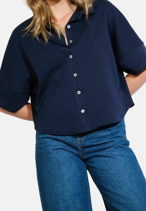Kvinde iført en løs mørkeblå skjorte med korte ærmer og blå højtaljede jeans mod en hvid baggrund.