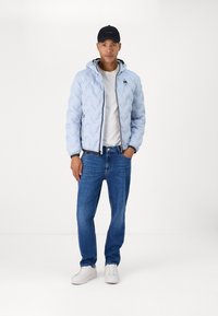 Giacca a vento azzurro chiaro con cuciture a zigzag, dettagli neri, cerniera frontale, indossata sopra una maglietta bianca e jeans blu, abbinata a sneakers bianche.