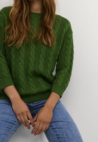 Grüner gestrickter Pullover mit Zopfmuster und 3/4-Ärmeln, kombiniert mit blauen Jeans. Der Pullover hat einen gerippten Saum am Halsabschluss und an den Ärmeln.
