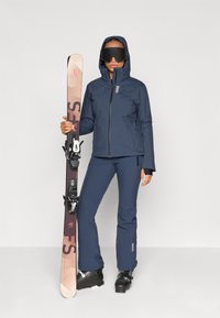 Ski oblečení zahrnující tmavě modrou voděodolnou bundu a kalhoty. Lyžař drží růžové a černé lyže s vázáním. Sněhule jsou černé.