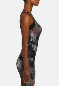 Kobieta w profilu bocznym, ubrana w bezrękawnik i legginsy z abstrakcyjnymi wzorami w czerni, bieli oraz stonowanych kolorach.