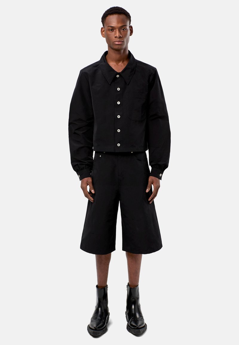 Svart kort jacka med stor krage, framknappar och en ficka, i kombination med knälånga svarta shorts och svarta ankelboots.