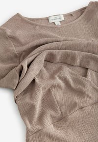 Haut de maternité en tissu texturé côtelé de couleur beige chaud, doté d'un col arrondi et d'un tombé souple avec des plis délicats.