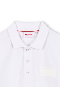 Polo blanc en tissu texturé, avec une patte de boutonnage à trois boutons, une finition rouge au col et un logo imprimé discret sur la gauche.
