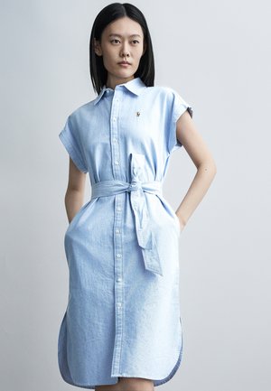 Kobieta stojąca z rękami w kieszeniach, ubrana w jasnoniebieską sukienkę koszulową z krótkim rękawem i paskiem na prostym jasnym tle.