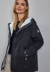 Veste imperméable noire avec doublure de capuche grise, fermeture éclair, deux poches latérales et détail de logo sur la manche. Tissu lisse avec une coupe décontractée.
