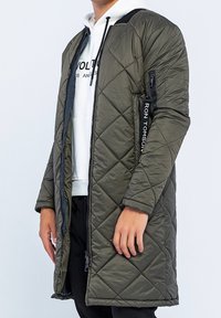 Olivengrøn lang quiltet frakke med lynlås og sidelommer, der har en sort accentstribet tag og en behagelig pasform.