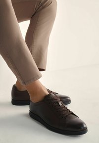 Brązowe skórzane sneakersy z okrągłym noskiem, sześcioma oczkami na sznurowadła i minimalistycznym designem. Czarna gumowa podeszwa i gładka faktura.