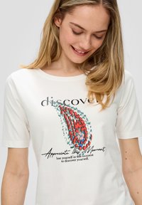 T-shirt blanc à manches courtes présentant un design graphique coloré avec des motifs rouges et bleus. Le texte indique "découvrez" et "Appréciez ce moment".