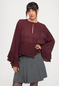 Blusa trasparente bordeaux con maniche lunghe a rouches sovrapposte e scollatura a chiave, abbinata a una gonna grigia plissettata.