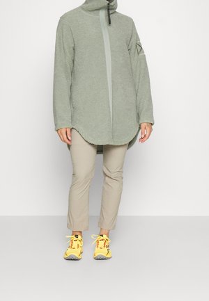 Persoon draagt een lichtgroen fleecejack met hoge kraag, beige broek en felgele sportschoenen, staand tegen een effen witte achtergrond.