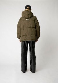 Olivgrüne Pufferjacke mit Kapuze, auf der Rückseite mit einem runden Logo versehen, kombiniert mit glänzenden schwarzen Lederhosen.