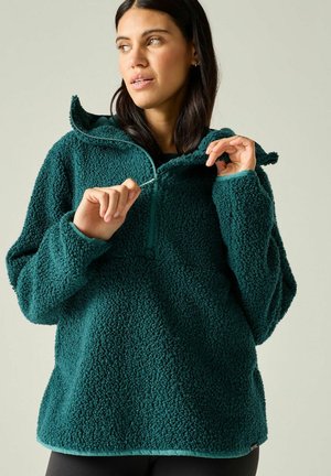 Mujer con cabello oscuro cerrándose una chaqueta de forro polar texturizada de color azul verdoso con cuello alto, sobre un fondo liso y claro.