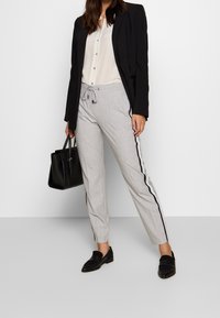 Lichtgrijze broek met een trekkoord in de taille, zwarte zijstrepen, gecombineerd met een zwart colbert en een zwarte handtas. Platte zwarte schoenen.