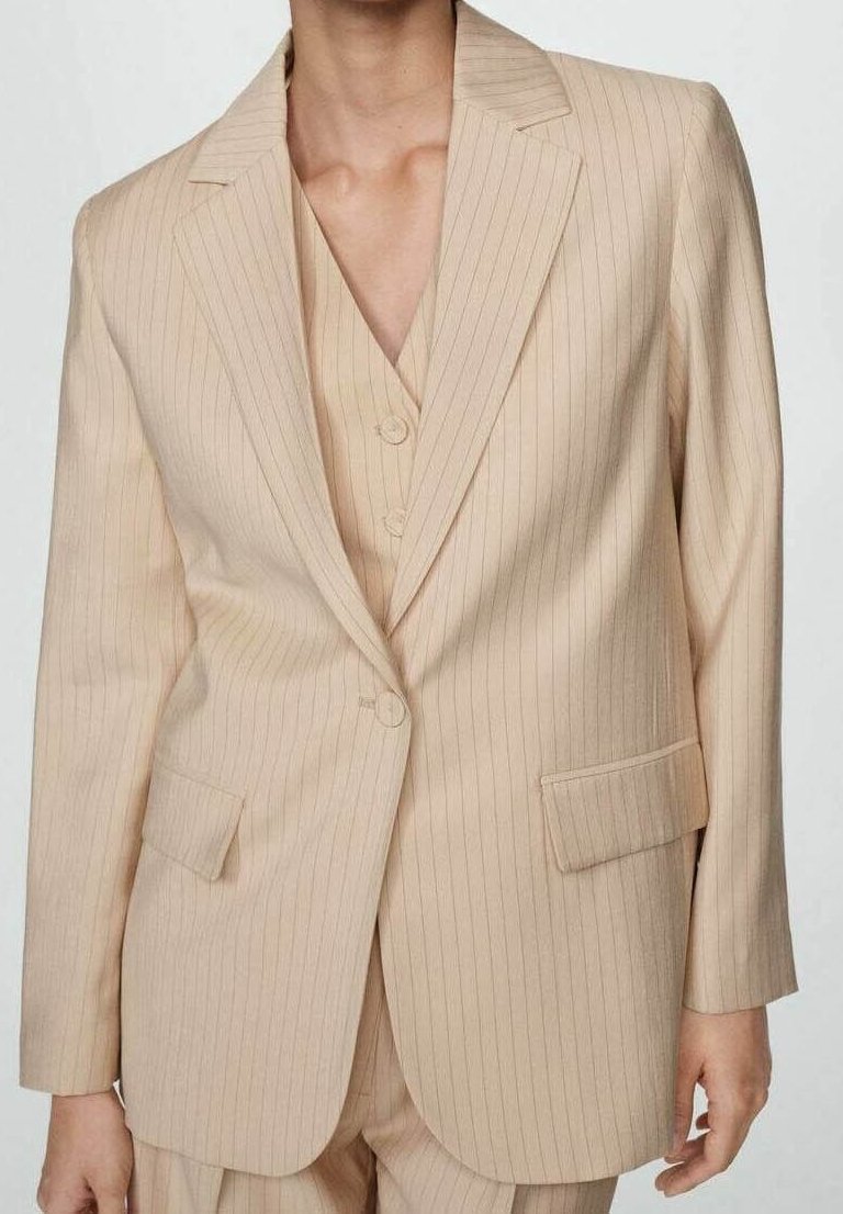 Femme portant un costume beige à fines rayures avec un blazer à un bouton et un gilet assorti boutonné en dessous.