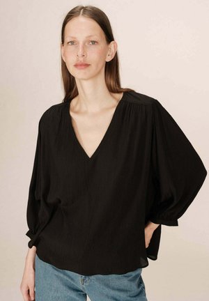 Blouse noire ample avec un décolleté en V et des manches bouffantes, en tissu texturé. L'ourlet est légèrement courbé. Associée à un jean bleu.