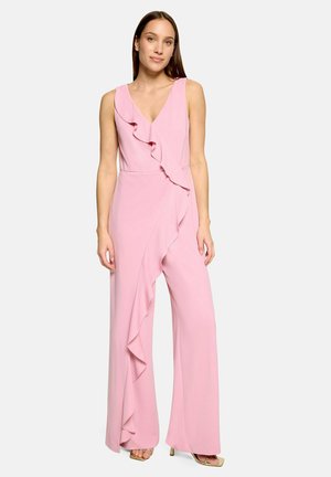 Femme portant une combinaison sans manches rose clair avec un col à volants et un devant froncé, debout les mains relâchées sur les côtés.