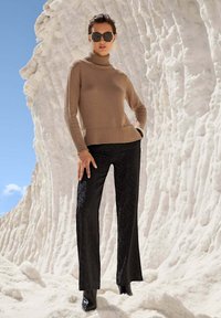 Beige coltrui met geribbelde details, gecombineerd met zwarte gestructureerde flared pants en zwarte hoge laarzen, tegen een zand-witte achtergrond.