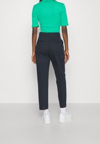 Donkerblauwe broek met een getailleerde pasvorm, voorzien van zijzakken en een gladde textuur. Gecombineerd met witte sneakers en een groene top.