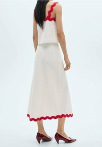 Vestido blanco con adornos ondulados rojos en los tirantes y el dobladillo. Textura suave, diseño sin mangas, llevado con zapatos de tacón rojo a juego.