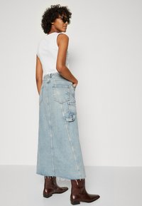 Lyseblå denimmaxiskjørt med frynset kant, sidelommer og en avslappet passform. Kombinert med brune ankelstøvler og en hvit ermeløs topp.