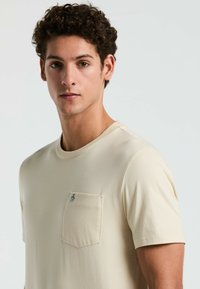 Beige T-shirt met korte mouwen, gemaakt van zachte stof, met een borstzak en een klein logoaccent. Eenvoudig, strak ontwerp met een ronde hals.