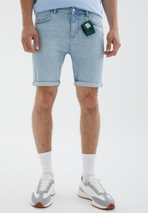Homme portant un short en denim bleu clair retroussé, des chaussettes blanches, des baskets gris et blanc, avec un porte-clés vert attaché à sa poche.