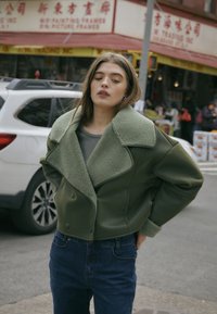 Olive green podwójnie zapinana kurtka z teksturowanym kołnierzem z owczej wełny, w połączeniu ze szarym topem i ciemnymi dżinsami.