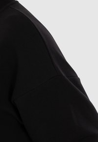 Gros plan sur la couture de l’épaule et de la manche d’un vêtement noir montrant la texture du tissu et les détails de la couture.