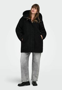 Črn, prenapihnjen plašč s kapuco, izdelan iz teksturirane tkanine; ima stranske žepke in sprednje zapiranje na gumb; oblečen čez sive široke hlače.