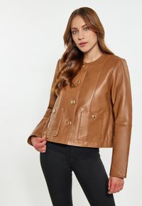 Brun læderjakke med rund halsudskæring, gyldne knapper, to forlommer og et tætsiddende design, parret med sorte bukser.