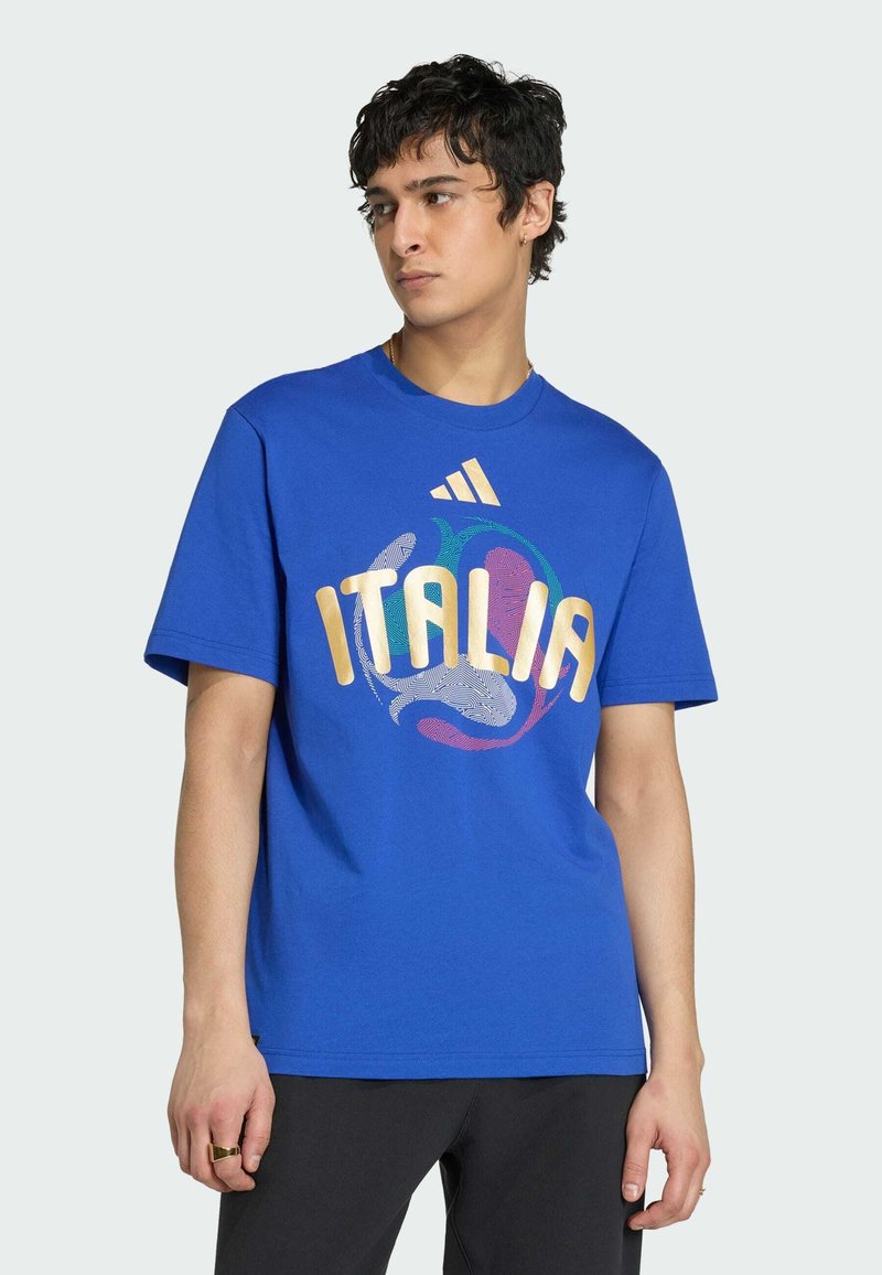 Jeune homme portant un T-shirt bleu avec le texte "ITALIA" et le logo Adidas, regardant sur le côté devant un fond clair uni.