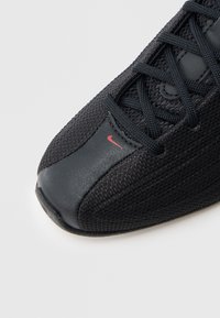 Czarny sportowy but z teksturowaną siateczkową górą, wyściełanym językiem i gładkim skórzanym noskiem, z małym czerwonym logo marki.