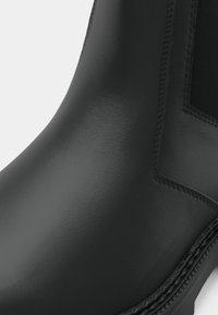 Κοντινό πλάνο από ένα λείο μαύρο δερμάτινο μποτάκι με λεπτομερές ράψιμο και ελαστική πλευρική πάνελ σε λευκό φόντο.