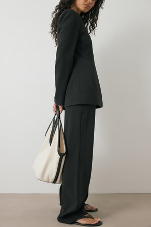 Black tailored suit with long sleeves, slim pants, and a black-and-white tote bag. Smooth texture, minimal design, and open-toed sandals.
