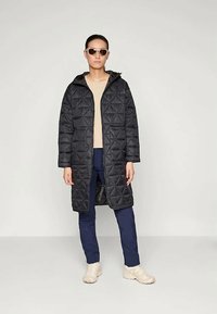 Melna dūku garā mēteli ar kapuci, kas ir ar ģeometrisku rakstu. Saskaņots ar bēša topu un zilganiem biksēm, pabeigts ar vieglām kedām.