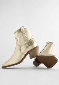 Botas de tobillo de cuero dorado con puntas afiladas, detalles ondulados y un tacón apilado. Presentan un lazo en la parte trasera y acentos de costura.