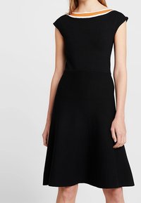 Žena v bezrukávových černých šatech se rozšířenou sukní a oranžovým a bílým lemováním kolem výstřihu.