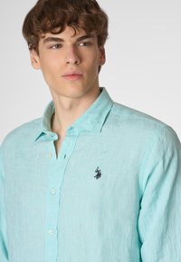 Camicia di lino turchese chiaro con colletto button-down, caratterizzata da un piccolo logo ricamato in nero sul lato sinistro del petto e tessuto strutturato.