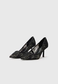 Pantofi cu toc înalt din dantelă neagră, cu vârf ascuțit, decorative cu modele florale și căptușeală neagră netedă, așezați pe un toc subțire.