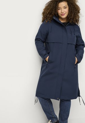 Kvinde med krøllet hår iført en mørkeblå lang frakke og jeans, hænderne i lommerne, stående mod en hvid baggrund.