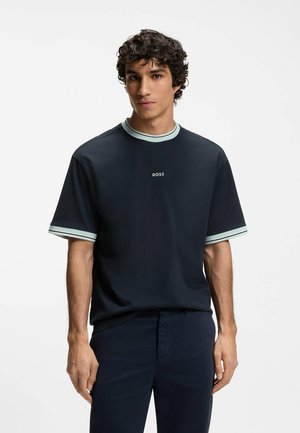 Junger Mann mit lockigem dunklem Haar, trägt ein marineblaues BOSS-T-Shirt mit hellblauen Abschlüssen am Kragen und den Ärmeln sowie dunkle Hosen.