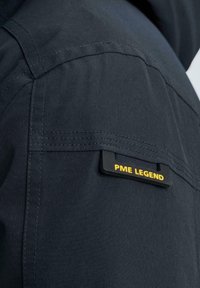 Giacca di tessuto blu navy con finitura testurizzata, cuciture visibili e un'etichetta nera con testo giallo "PME LEGEND" applicata sulla spalla.