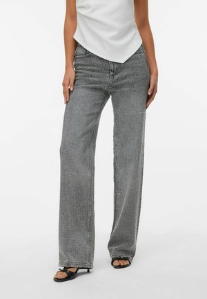 Vrouw in grijze wijde broek met een wit stippenpatroon, een witte asymmetrische top en zwarte open hakken op een witte achtergrond.