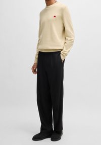Pull beige en maille avec col rond et petit détail de logo rouge, associé à un pantalon noir à jambes larges et des baskets noires. Texture lisse.
