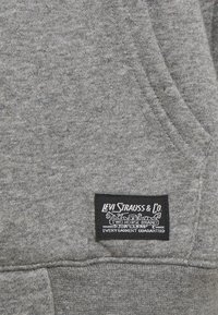 Grå sweatshirt med en mjuk textur, som har en svart etikett med vit text och illustration i det nedre hörnet. Enkelt design.